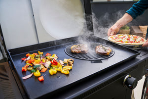 A Char-Broil grill with steaks, grilled vegetables, and a person placing a pizza on the cooking surface.
