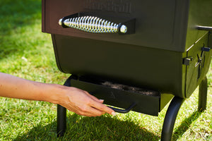 Hand pulling out the ash pan from a Char-Broil charcoal grill for easy cleaning on a grassy lawn.