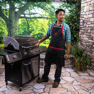 Person in apron stands beside a Char-Broil gas grill on a patio, ready for outdoor grilling. Lush greenery in background.