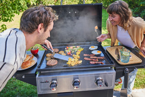 Two people grilling burgers, eggs, bacon, and veggies on a Char-Broil gas griddle outdoors, with buns and drinks nearby.