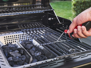 Hand cleaning grill grates with a brush; charcoal and warming rack visible inside Char-Broil grill.