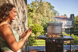 Person using a smartphone to monitor a Char-Broil smart grill cooking food on a sunny patio.