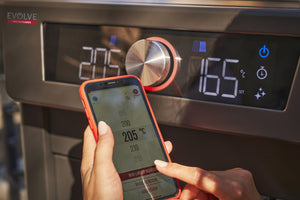 Close-up of a person adjusting a Char-Broil EVOLVE grill temperature using a smartphone app and digital control panel.