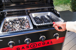 Close-up of a Char-Broil Gas2Coal grill with charcoal trays, showing both fresh and used charcoal. Hand holding grill tool.